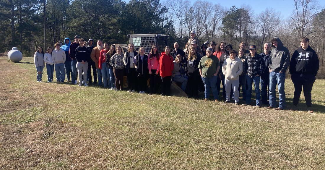 Students and instructors at Cleburne County Career Technical School stand in a grassy area in front of a skid steer on a sunny day.